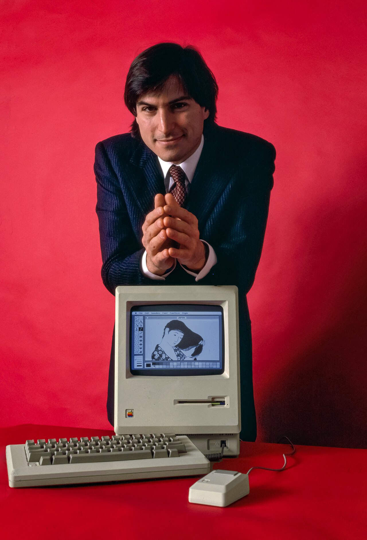 Homem jovem de terno azul-escuro e gravata, com as mãos unidas, apoiado atrás de um Macintosh original com teclado e mouse, sobre fundo vermelho.
