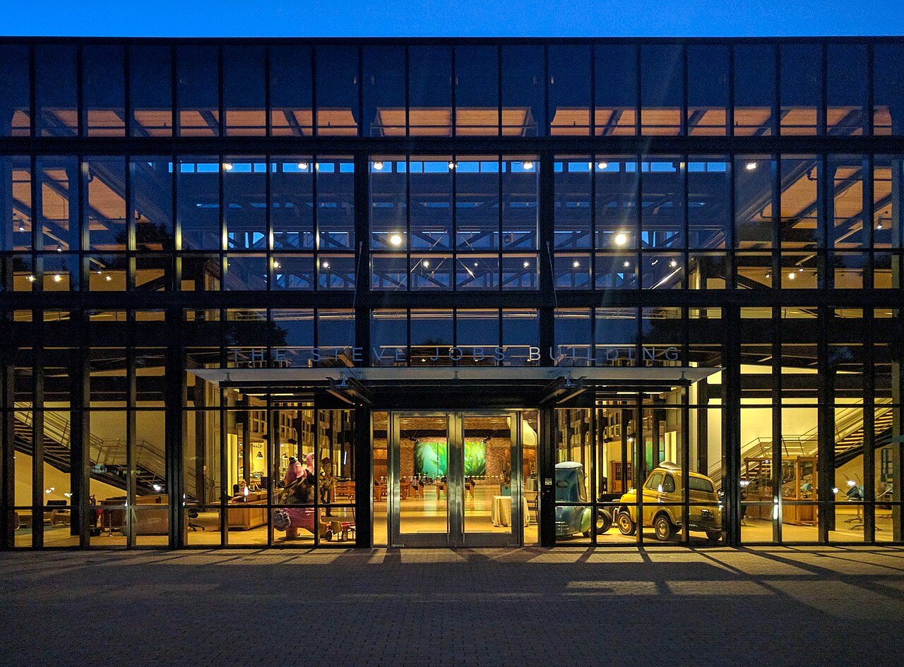 Edifício moderno de vidro e tijolos ao entardecer, com as janelas iluminadas: o prédio da Pixar que leva o nome de Steve Jobs.