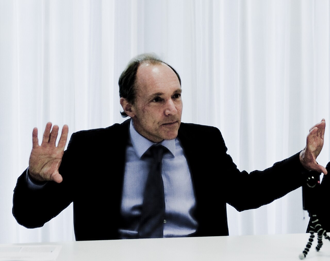 Tim Berners-Lee de terno escuro, camisa azul e gravata, sentado diante de cortinas brancas, com as duas mãos abertas ao falar.