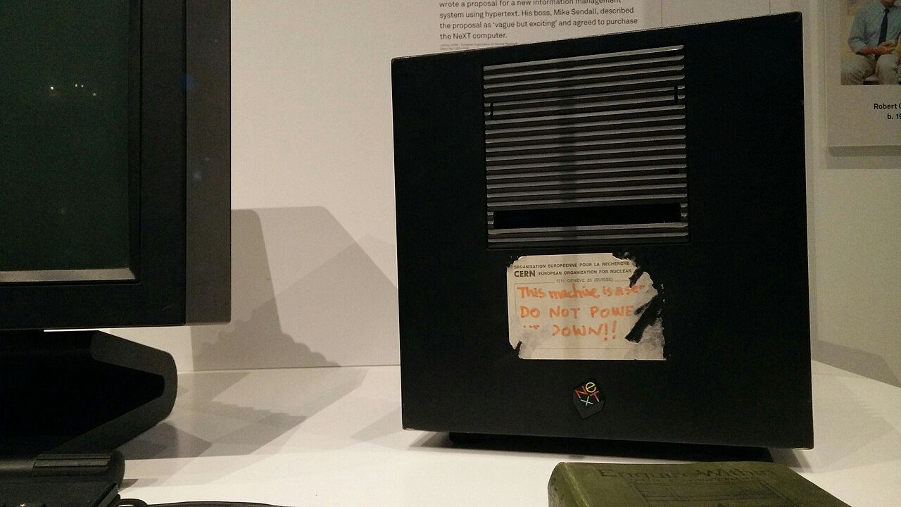 Detalhe de um computador NeXT preto com uma etiqueta de papel escrita à mão em laranja, num museu, ao lado de um monitor desligado.