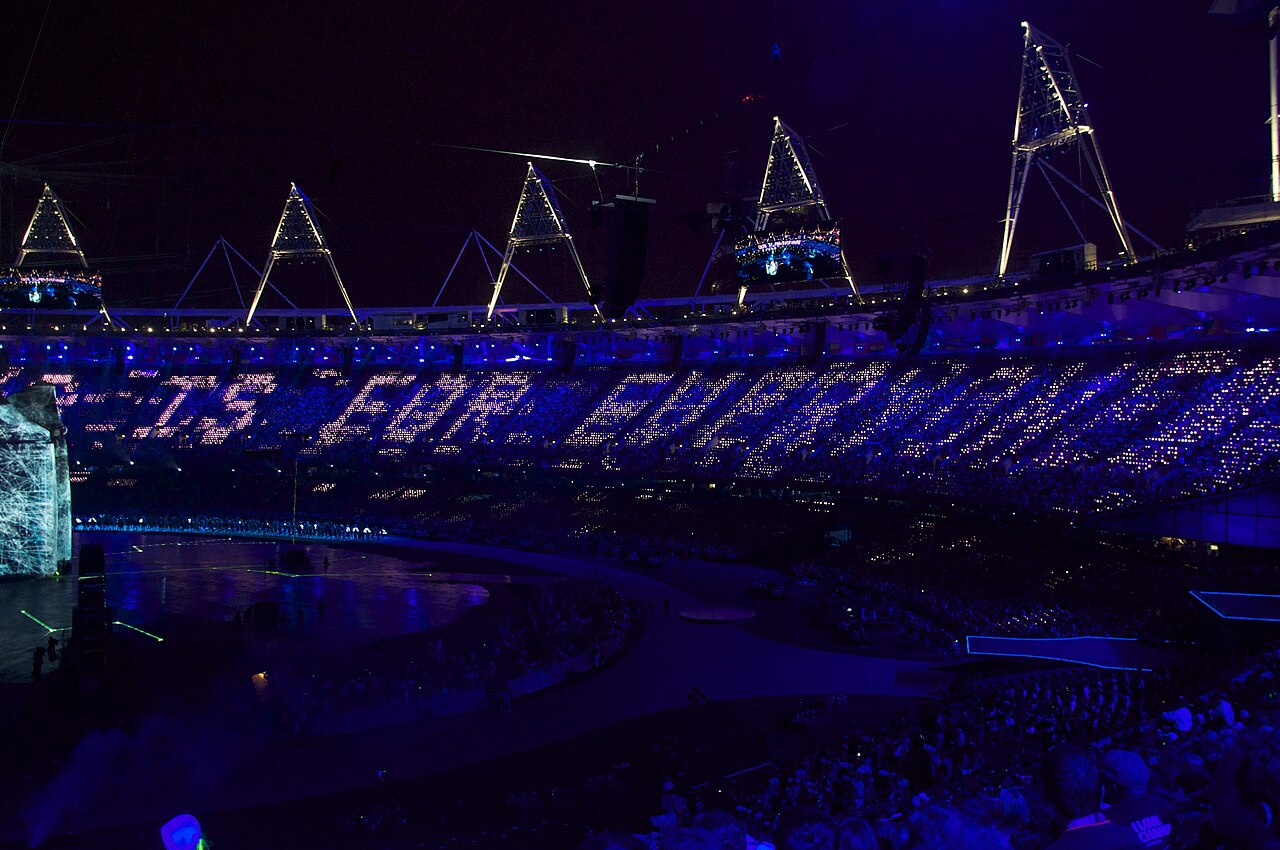 Arquibancadas de um estádio à noite, em azul e roxo, com milhares de luzes formando letras luminosas nas cadeiras.