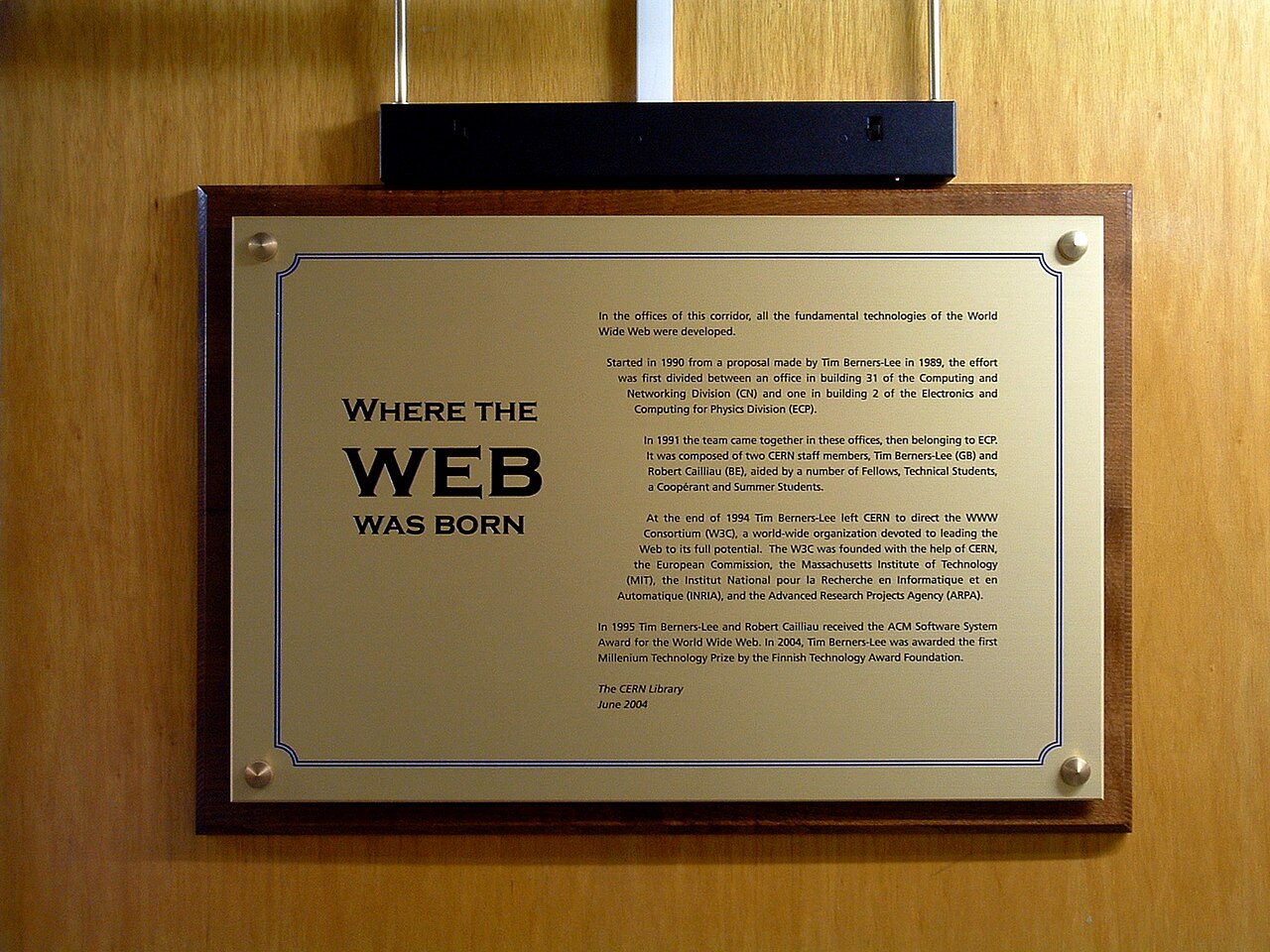 Placa dourada numa porta de madeira, com o título "Where the WEB was born" e um texto em inglês sobre o grupo do CERN e o W3C.