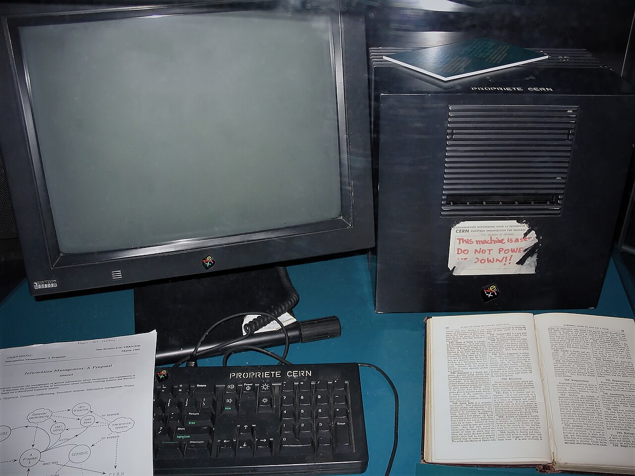 Computador NeXT preto com monitor, teclado e etiqueta escrita à mão em vermelho, numa vitrine, com uma cópia impressa da proposta ao lado.