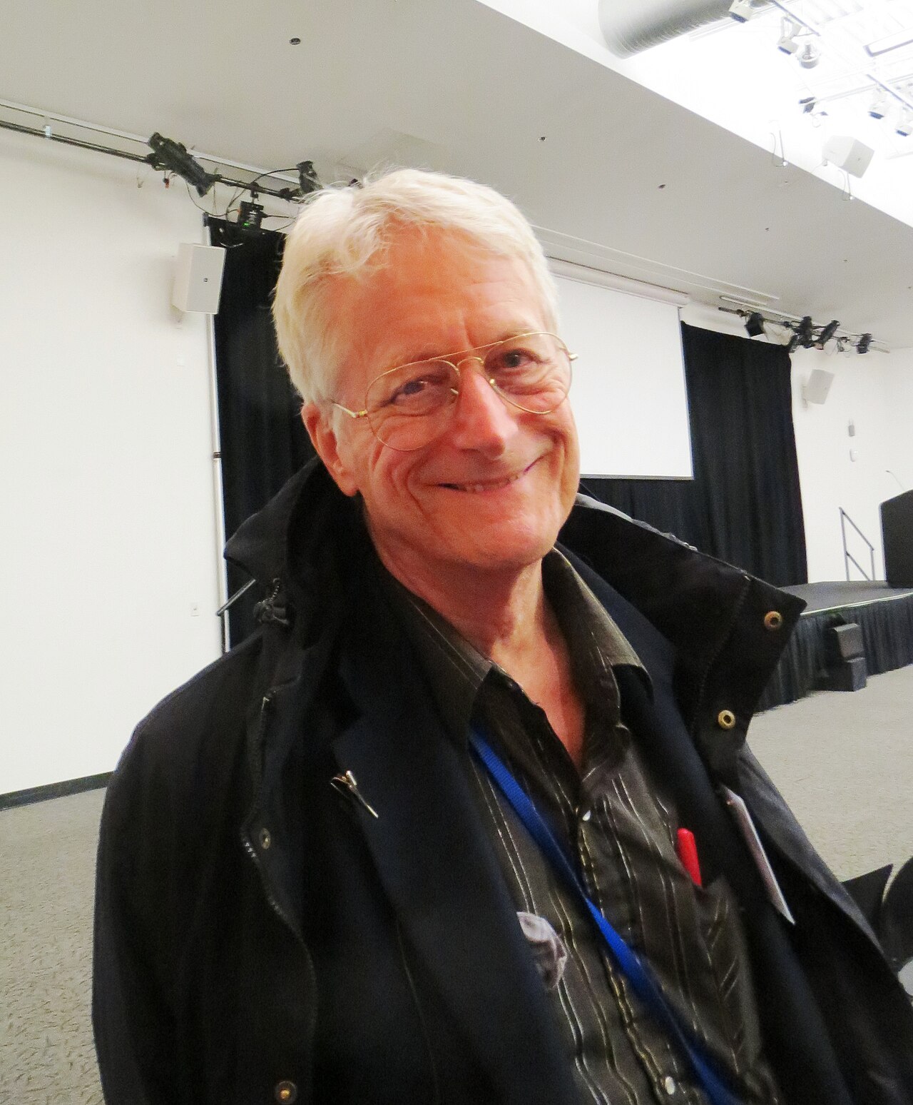 Ted Nelson, de cabelos brancos e óculos de aro fino, sorrindo para a câmera com um casaco escuro, num salão de eventos.