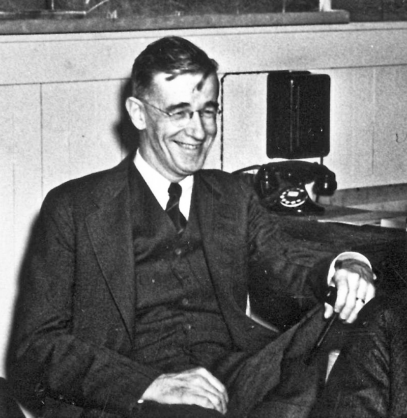Foto em preto e branco de Vannevar Bush, de óculos, terno e gravata, sorrindo sentado numa sala com um telefone na parede.